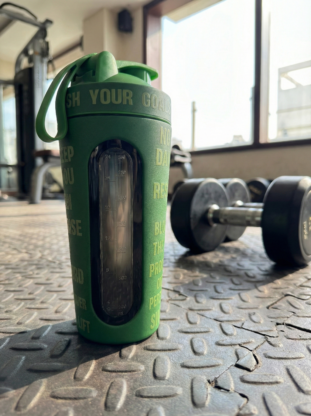 Green BEASTOX Performance Shaker placed on gym floor with dumbbells, showcasing its rugged design and clear measurement window.